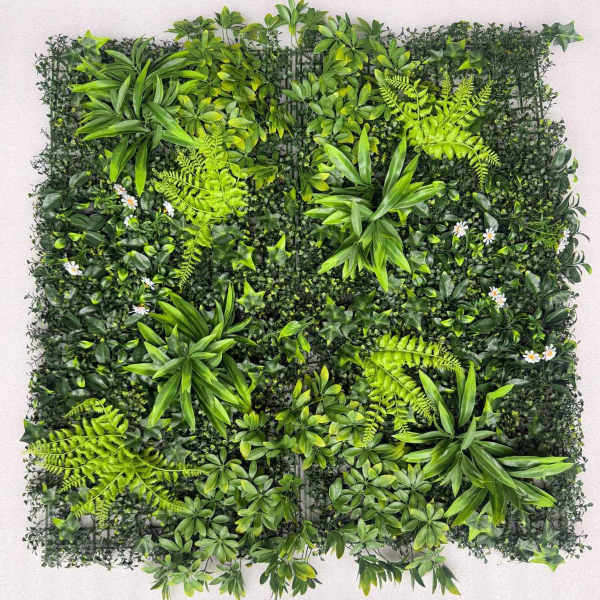A square artificial greenery wall panel featuring dense faux foliage, ferns, small white flowers, and leafy vines mounted on a black wire grid frame, shown against a light neutral background.