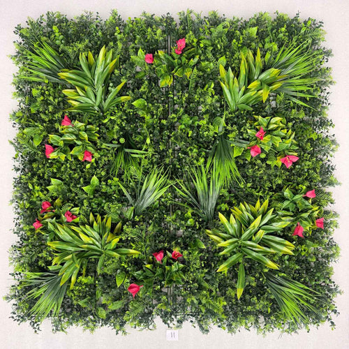 Square artificial green wall panel featuring dense faux foliage, palm-like leaves, and scattered bright pink flowers, photographed against a plain light background.