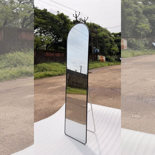 A full-length arched floor mirror with a slim black frame and a metal tripod stand, set outdoors on a white sheet of paper.