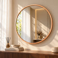 A large round wall mirror with a warm copper-toned frame hangs on a beige wall above a wooden console table. The mirror reflects a softly lit interior with curtains and a vase, while the room itself is styled with a reed diffuser, books, and a bowl.