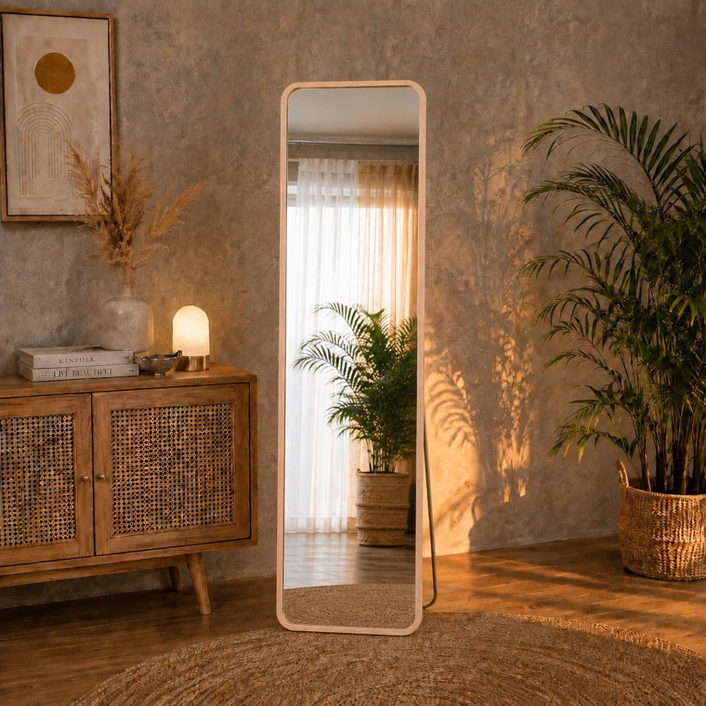 A tall, full-length standing mirror with a light aluminum alloy frame stands in a warm, boho-style room. The standing mirror reflects a soft light from a curtain in the background